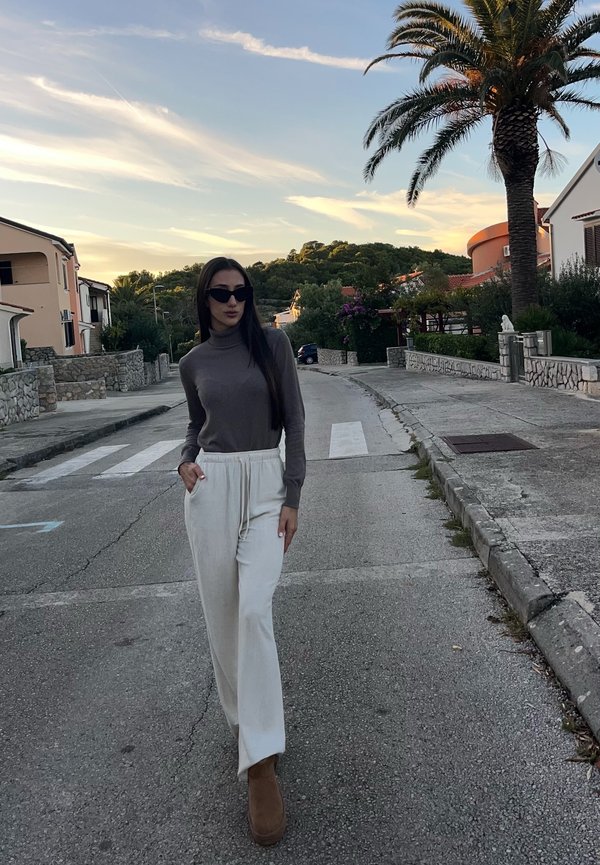 Une femme portant des lunettes de soleil, un haut gris, un pantalon blanc et des bottes brunes marche dans une rue résidentielle calme au coucher du soleil.