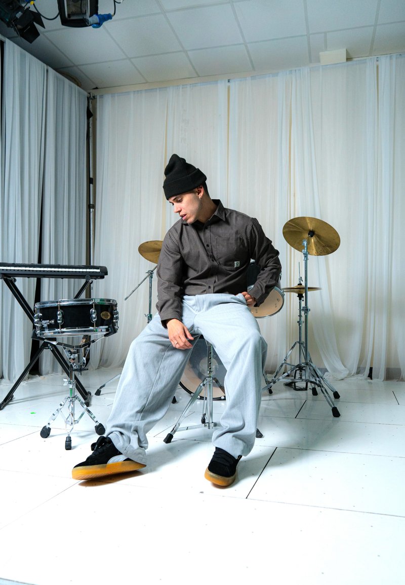 Beanie nero, camicia a maniche lunghe a quadri marroni, pantaloni grigi a gamba larga e sneakers nere con suole color gomma. Batteria e tastiera sullo sfondo.