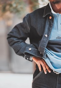 Denim jacket with contrasting stitching and buttons, layered over a blue polo shirt. Dark blue jeans featuring a visible belt and textured fabric.