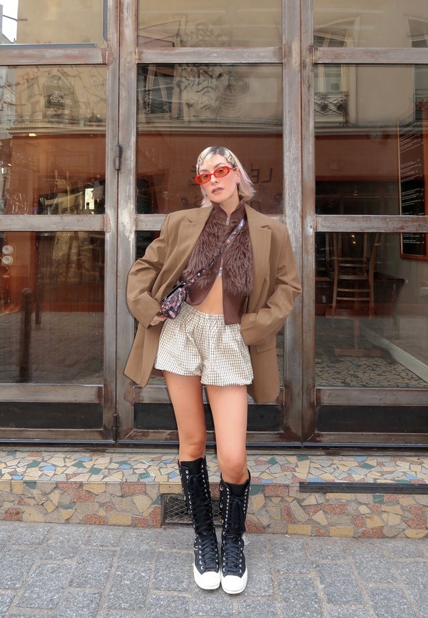 Donna con capelli biondi corti che indossa occhiali da sole arancioni, blazer marrone oversize, top peloso, pantaloncini a quadri e stivali neri con lacci, in piedi in strada.