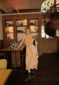 Oranje en crèmekleurige jurk met een vloeiende snit, gecombineerd met zwarte combatlaarzen. Accessoires omvatten een kettingriem en een kleurrijke tas.