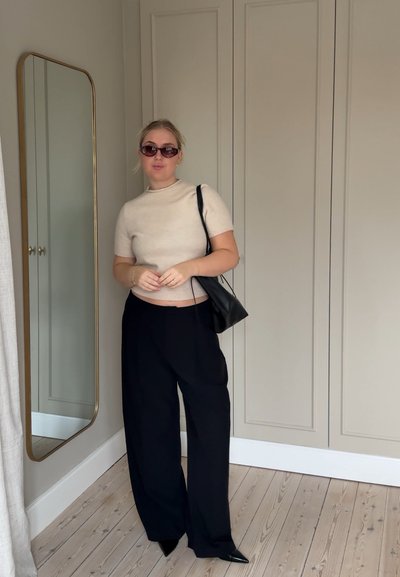 Suéter de manga corta beige, pantalones de pierna ancha negros, bolso negro y gafas de sol oscuras. Un espejo de cuerpo entero refleja el atuendo. Suelo de madera clara.