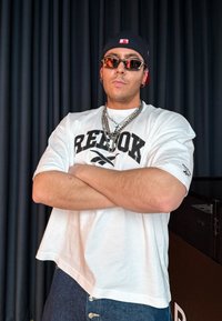 Άνδρας που φοράει λευκό πουκάμισο Reebok, αλυσίδες ασημένιες σε στρώματα, γυαλιά ηλίου και μαύρο καπέλο αντίστροφα, στέκεται με τα χέρια σταυρωμένα μπροστά σε σκοτεινό φόντο.