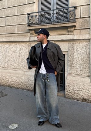 Hombre con abrigo gris oscuro, gorra negra, jeans holgados y zapatos negros, sosteniendo una bolsa negra y usando auriculares en una acera de la ciudad.