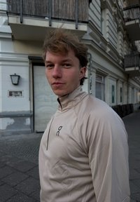 Fiatal férfi világosbarna hajjal, bézs színű, negyed cipzáras sportfelsőt viselve, egy városi járdán áll, közel lakóépületekhez.