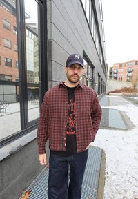 Červená a čierna kockovaná košeľa na gombíky cez čierne tričko s potlačou. Modré džínsy z denimu, stojaci v exteriéri v mestskom prostredí pri odrazivých oknách.