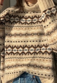 Femme portant un pull à col roulé en maille beige avec des motifs géométriques marron, noirs et blancs, associé à un jean en denim bleu.