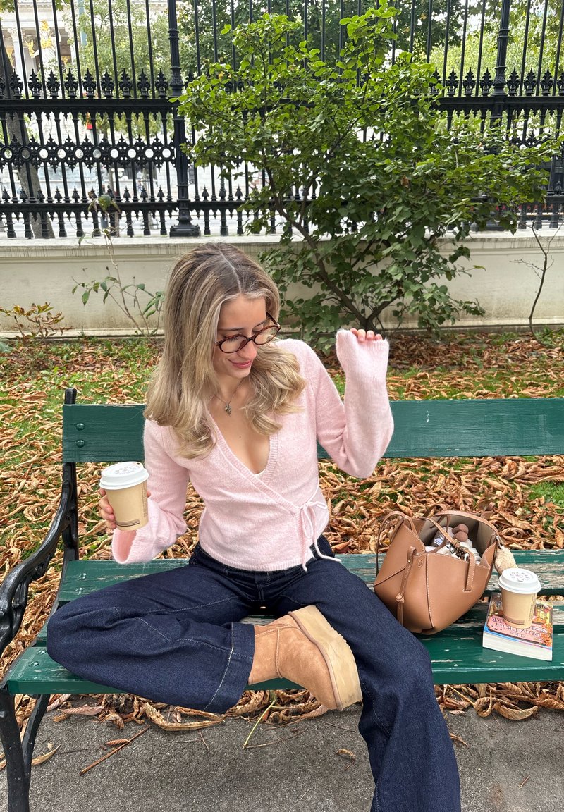 Light pink, wrap-style sweater with bell sleeves; denim pants; tan suede boots; brown handbag; disposable coffee cup; autumn leaves on ground.