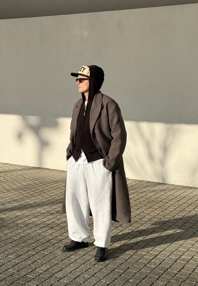Persona con un abrigo de cuadros, sudadera negra con capucha, gorra blanca, gafas de sol, pantalones deportivos grises y zapatos negros, de pie sobre una superficie pavimentada al aire libre.