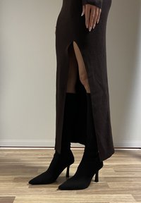 Femme portant une jupe côtelée marron foncé fendue à mi-cuisse et des bottines à talons noires à bout pointu, debout sur un sol en bois.