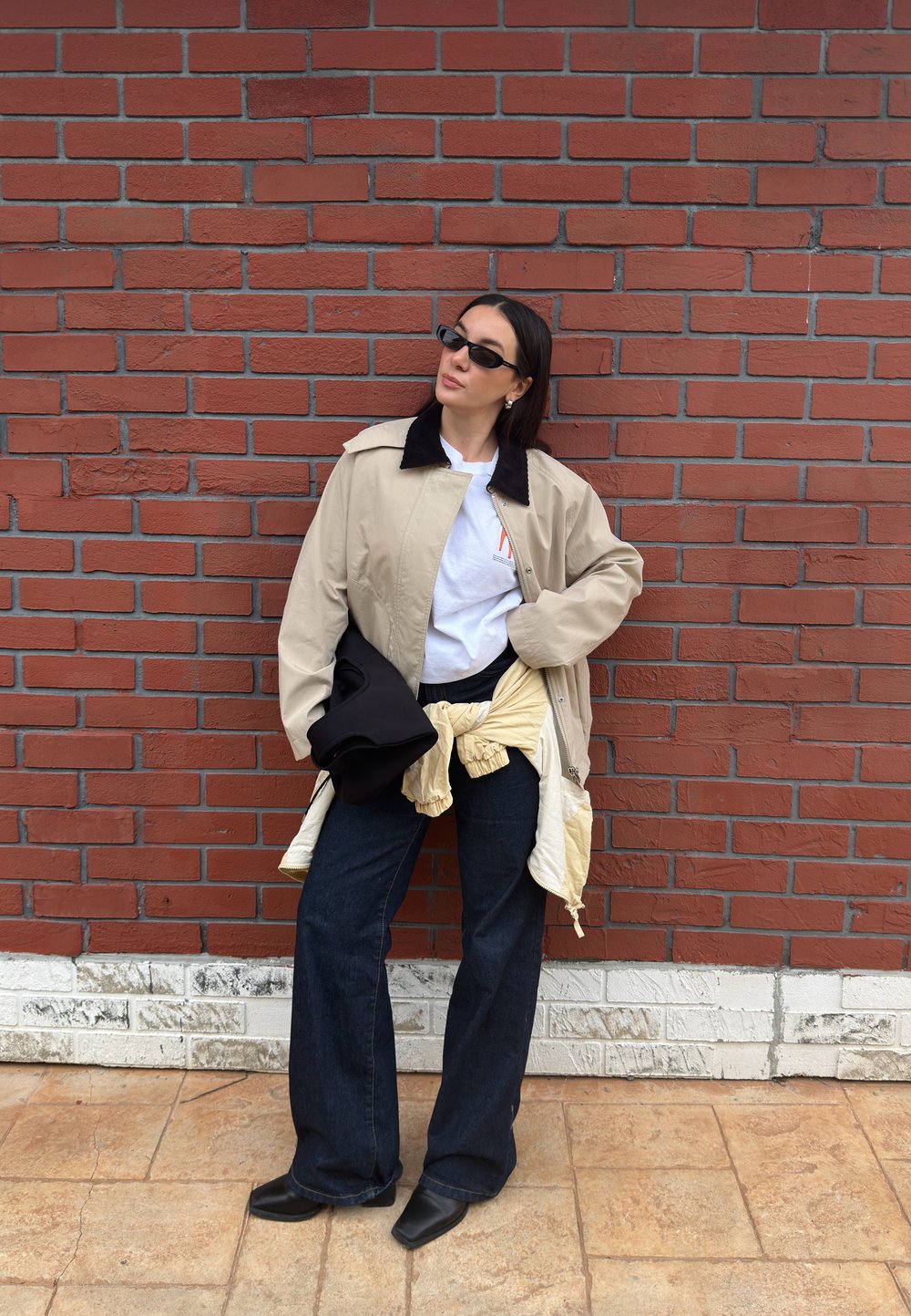 Femme portant une veste beige, une chemise blanche, un jean foncé, des lunettes de soleil noires et des chaussures noires, debout contre un mur en briques rouges, une main sur la hanche.