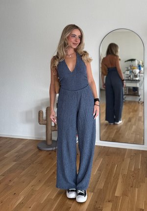 Mono gris canaleado con escote en V profundo y pantalones anchos, combinado con zapatillas negras. El fondo incluye una decoración de cactus y un espejo.