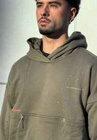 Sweat à capuche vert olive avec une poche zippée à l'avant, arborant un texte réfléchissant "run with the mind" et des motifs discrets en points. Tissu en mélange de coton doux.