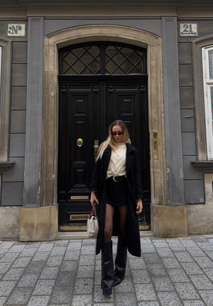 Mujer con abrigo negro, botas, suéter beige, gafas de sol y bolso beige caminando por una calle empedrada frente a unas puertas dobles negras.