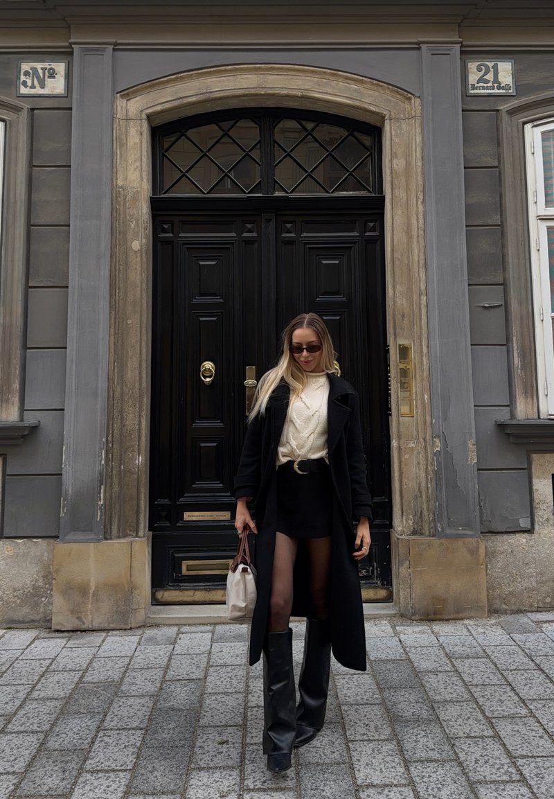 Femme portant un manteau noir, des bottes, un pull beige, des lunettes de soleil, tenant un sac à main beige, marchant sur une rue pavée devant des portes doubles noires.
