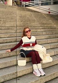 Un vestă albă tricotată cu dungi roșii și albastre peste un tricou cu mâneci lungi bordo, asortată cu o fustă cu dungi subțiri. Geantă albă și adidași robusti.