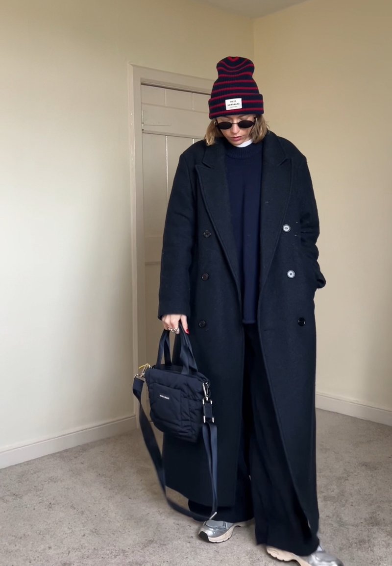 Person trägt einen langen schwarzen Mantel, schwarze Hose, marineblauen Pullover, gestreiften Beanie, Sonnenbrille und silberne Turnschuhe und hält eine schwarze Handtasche drinnen.