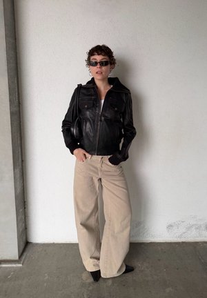 Persona con chaqueta de cuero negra, pantalones anchos beige, zapatos negros puntiagudos, gafas de sol oscuras, de pie contra una pared blanca con las manos en los bolsillos.
