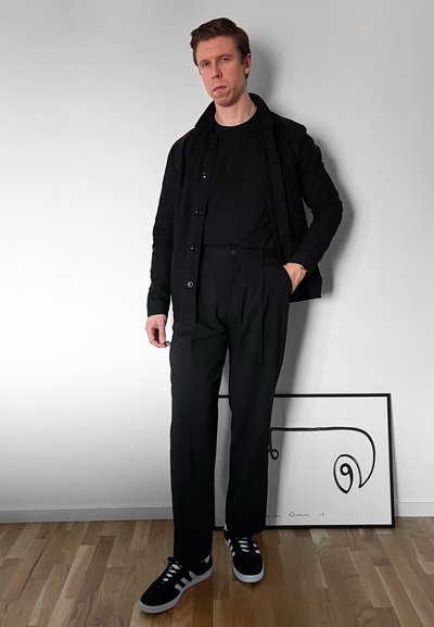 Chaqueta negra, camiseta negra y pantalones plisados negros, combinados con zapatillas negras con detalles en blanco; de pie frente a un fondo claro.