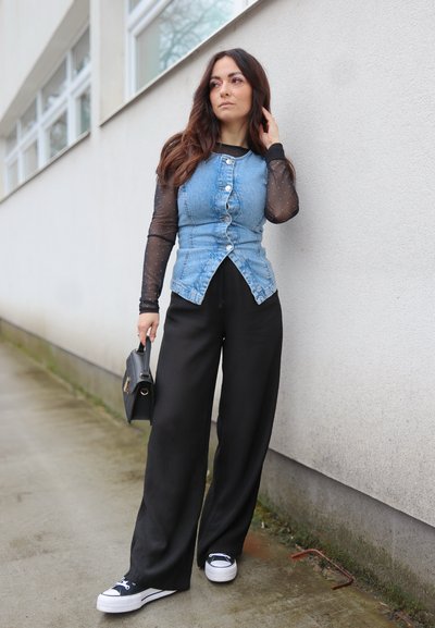 Chaleco de mezclilla en azul claro con botones delanteros, combinado con una blusa negra de manga larga y transparente. Pantalones negros holgados y zapatillas negras. Bolso negro.