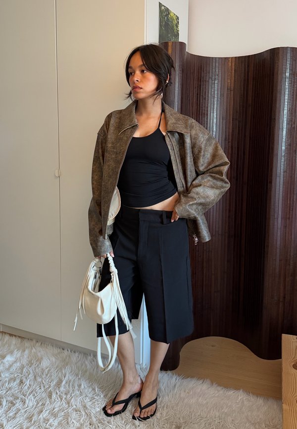 Femme portant une veste marron surdimensionnée, un débardeur noir, un short long noir et des sandales noires, tenant un sac blanc à l'intérieur.