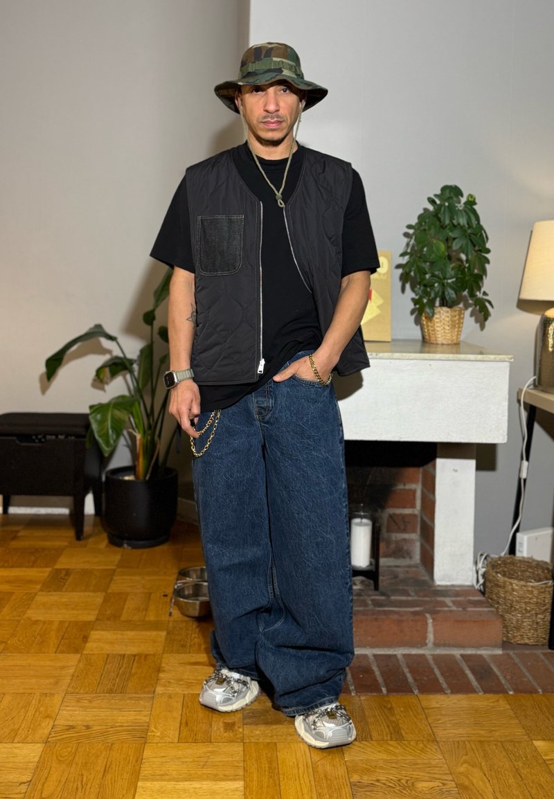 Mand iført camouflage bøllehat, sort vest, store blå jeans og hvide sneakers står indenfor ved planter og en pejs.