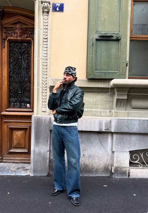 Un homme portant une veste en cuir noire, un bonnet à motifs et un jean ample se tient pensif à côté d’une entrée ornée, dans une rue de la ville.