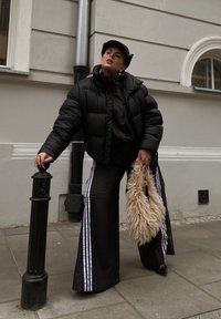Casaco puffer preto, blusa preta, calças flare pretas com riscas brancas; segurando uma mala de mão fofinha bege; com um fundo cinza claro.