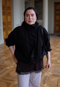 Coupe-vent noir surdimensionné à col montant, superposé à un haut marron transparent orné de dentelle, associé à un pantalon de couleur claire.