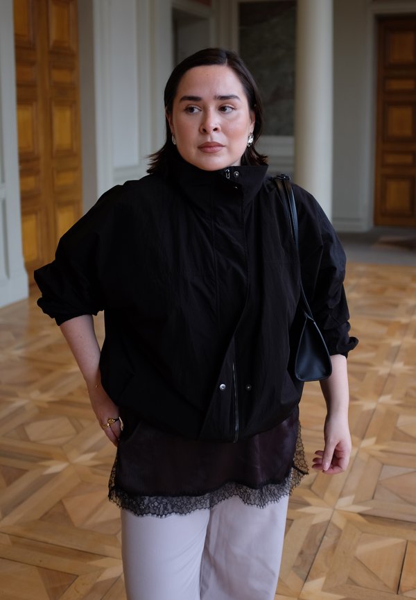 Antivento nero oversize con collo alto, abbinato a una maglia marrone trasparente con finiture in pizzo e pantaloni chiari.