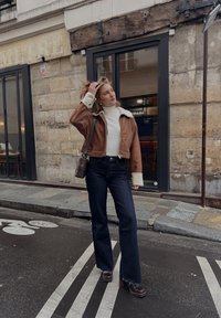 Bruine cropped jack met schapenvachtkraag, witte turtleneck, hoge taille donkere jeans en bruine hakken, afgebeeld tegen een stenen achtergrond.