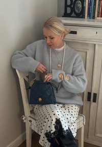 Femme en pull gris et jupe blanche à pois assise sur une chaise, tenant un sac matelassé bleu marine avec une bandoulière chaîne dorée.