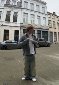 Καρό πουκάμισο με μακριά μανίκια πάνω από λευκό t-shirt, συνδυασμένο με ανοιχτό μπλέ τζιν με λεκέδες και τρύπες. Λευκά αθλητικά παπούτσια ολοκληρώνουν την εμφάνιση.