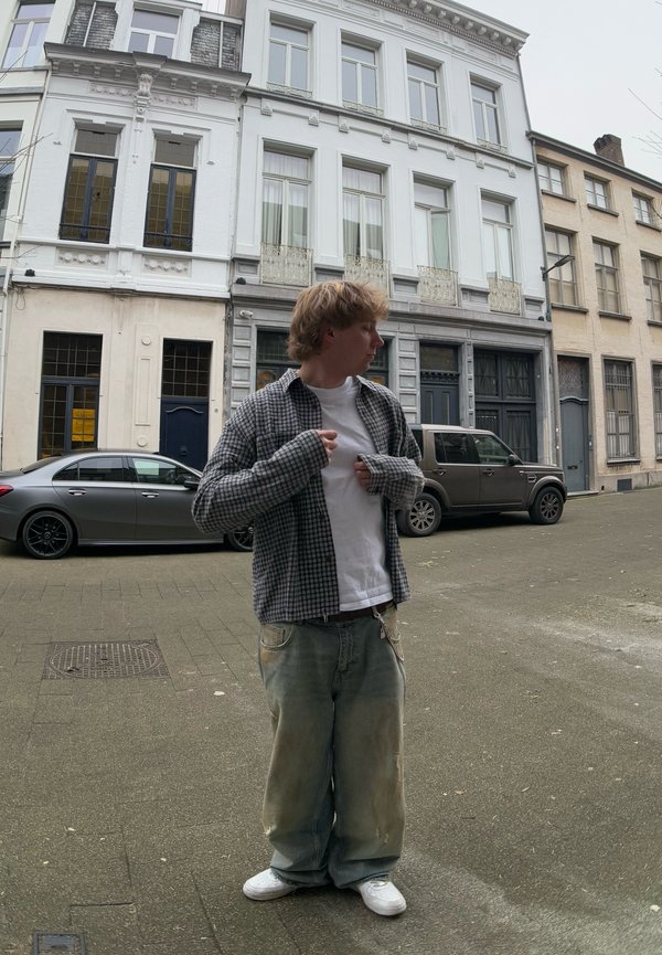 Karirana srajca z dolgimi rokavi čez belo majico, v kombinaciji z svetlo modrimi jeans hlačami, ki imajo madeže in solze. Bele superge zaokrožijo videz.