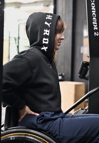 Personne en sweat à capuche noir assise dans un fauteuil roulant, mains sur les hanches, tournée vers la droite, près d'un rameur Concept2 dans une salle de sport.