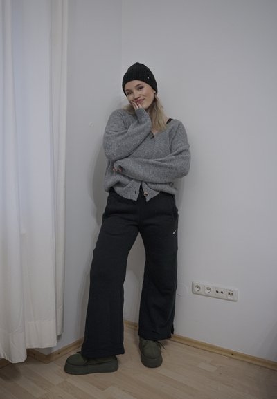 Cardigan de punto gris con botones, pantalones de chándal negros y zapatos verdes. De pie contra una pared lisa, con cortinas blancas y suelo de madera.