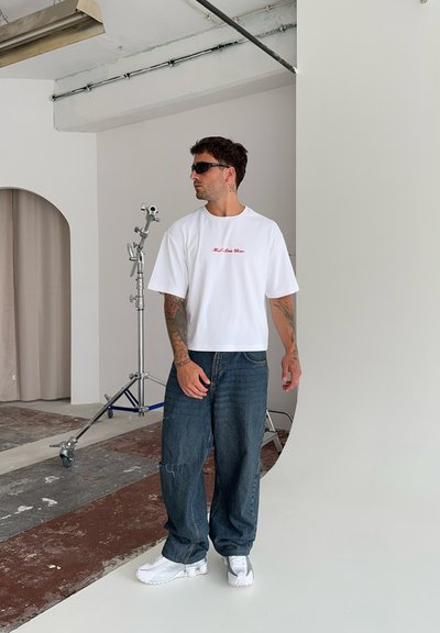 Camiseta blanca corta con texto rojo, combinada con pantalones de mezclilla azul desgastados y zapatillas blancas; modelo usando gafas de sol. Fondo de estudio.