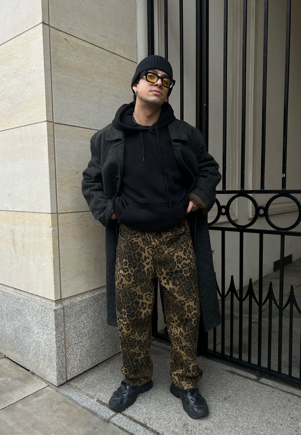 Siv volnen plašč čez črn kapuco, v kombinaciji z rjavimi hlačami z leopardjim vzorcem in črnimi športnimi čevlji. Dopolnjen z rumenimi sončnimi očali in kapo.