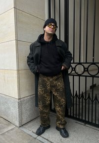 Siv volnen plašč čez črn kapuco, v kombinaciji z rjavimi hlačami z leopardjim vzorcem in črnimi športnimi čevlji. Dopolnjen z rumenimi sončnimi očali in kapo.