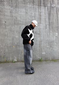 Juodai baltas karvės rašto pūkų džemperis su aukštu apykakle, derinamas su laisvomis pilkomis kelnėmis ir juodomis šlepetėmis ant tekstūruoto betono fono.