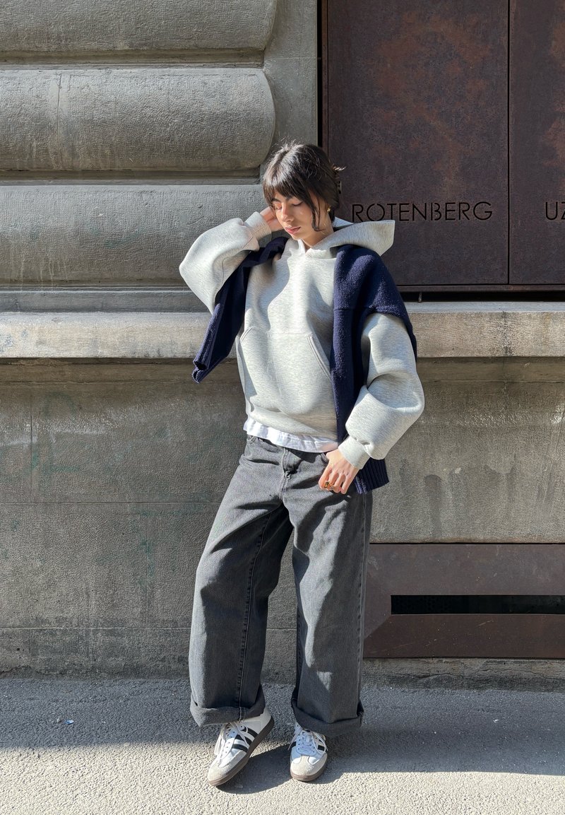 Jeune personne en sweat à capuche gris et jeans larges, appuyée contre un mur en pierre, avec un pull bleu marine sur les épaules, regardant pensivement vers le bas.