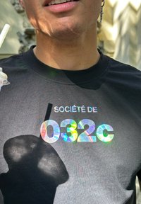 T-shirt noir arborant "SOCIÉTÉ DE" en blanc au-dessus du texte brillant et holographique "032c." Le tissu semble lisse avec un col rond classique.