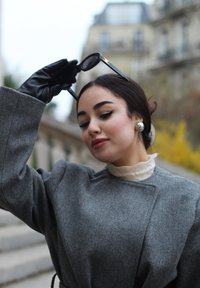 Grahamfarbener Wollmantel mit weiten Ärmeln, weißem Spitzenkragen, schwarzen Lederhandschuhen und dunkler Sonnenbrille, die in einer Hand gehalten wird. Insgesamt glatte Textur.