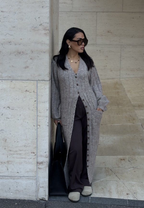 Langer, grauer Strickcardigan mit dunklen Knöpfen, seitlichen Taschen und geripptem Muster, kombiniert mit braunen Hosen und cremefarbenen Hausschuhen, während eine schwarze Tasche gehalten wird.