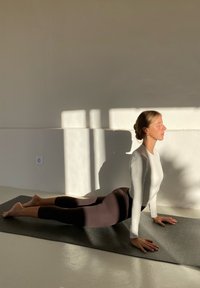 Yogastilling på grå måtte; person iført en tætsiddende hvid langærmet trøje og brune leggings; blød belysning kaster skygger på en hvid væg.