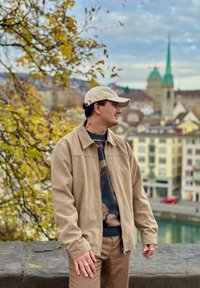 Beige Wildlederjacke über einem Pullover mit Tarnmuster, kombiniert mit braunen Hosen. Das Modell trägt eine cremefarbene Mütze und silberne Ringe.