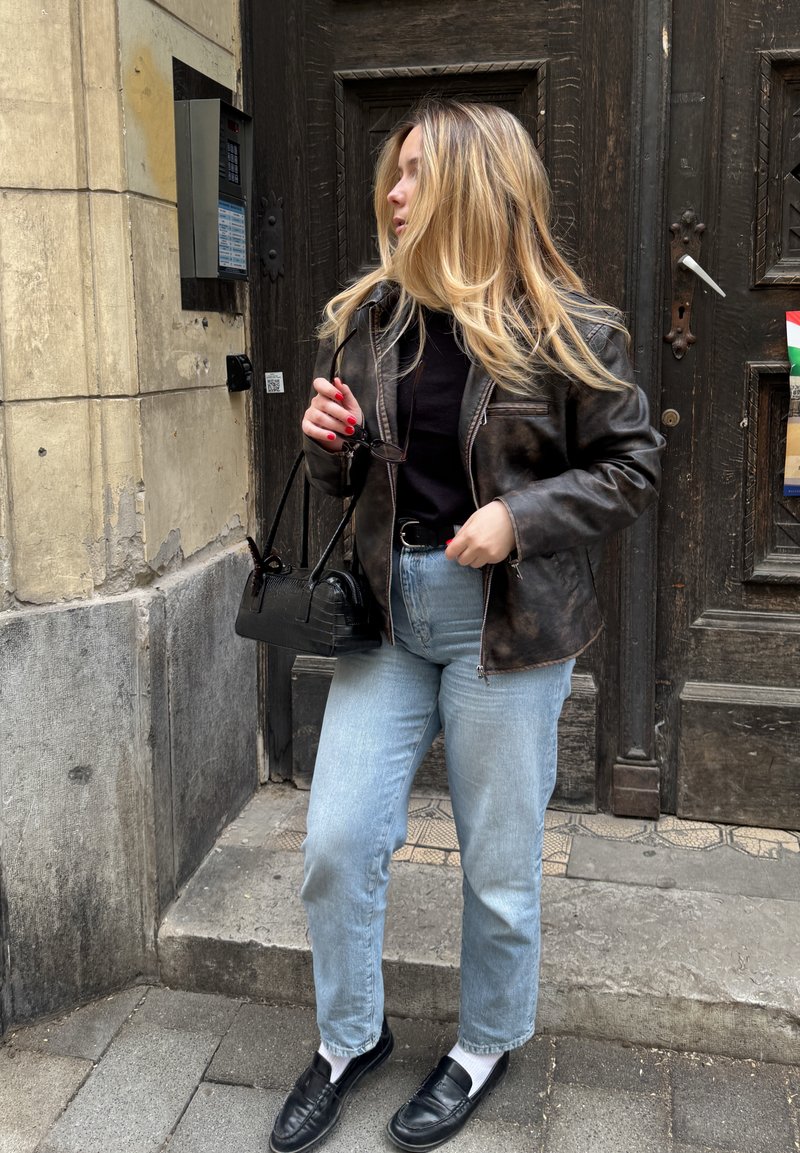 Femme blonde avec veste en cuir et jean clair se tient sur une marche en pierre devant une porte en bois sombre, tenant un petit sac à main noir et des lunettes.