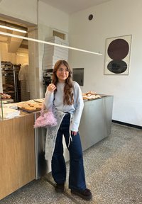 Maglione grigio con bottoni, jeans blu scuro a gamba larga, borsa rosa pelosa, in piedi accanto a una vetrina di paste in una pasticceria con arredamento dai toni neutri.