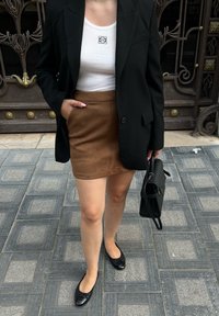 Μαύρο σακάκι πάνω από λευκό ραφιναρισμένο τοπ, καφέ σουέτ μίνι φούστα, μαύρα φλάντς με λεπτομέρεια φιόγκου, κρατώντας μια μαύρη δομημένη τσάντα.