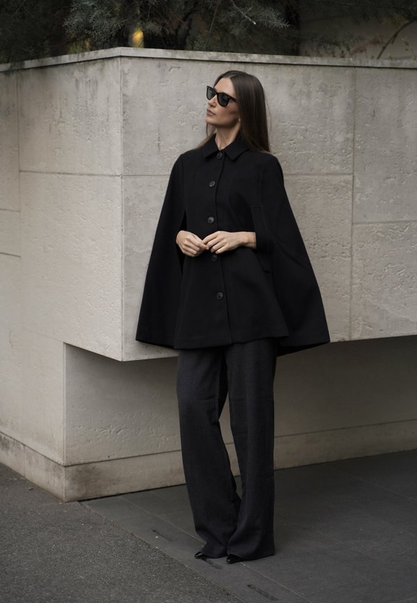 Mujer con capa negra abotonada, pantalones anchos oscuros y gafas de sol de pie contra una pared beige con textura al aire libre.